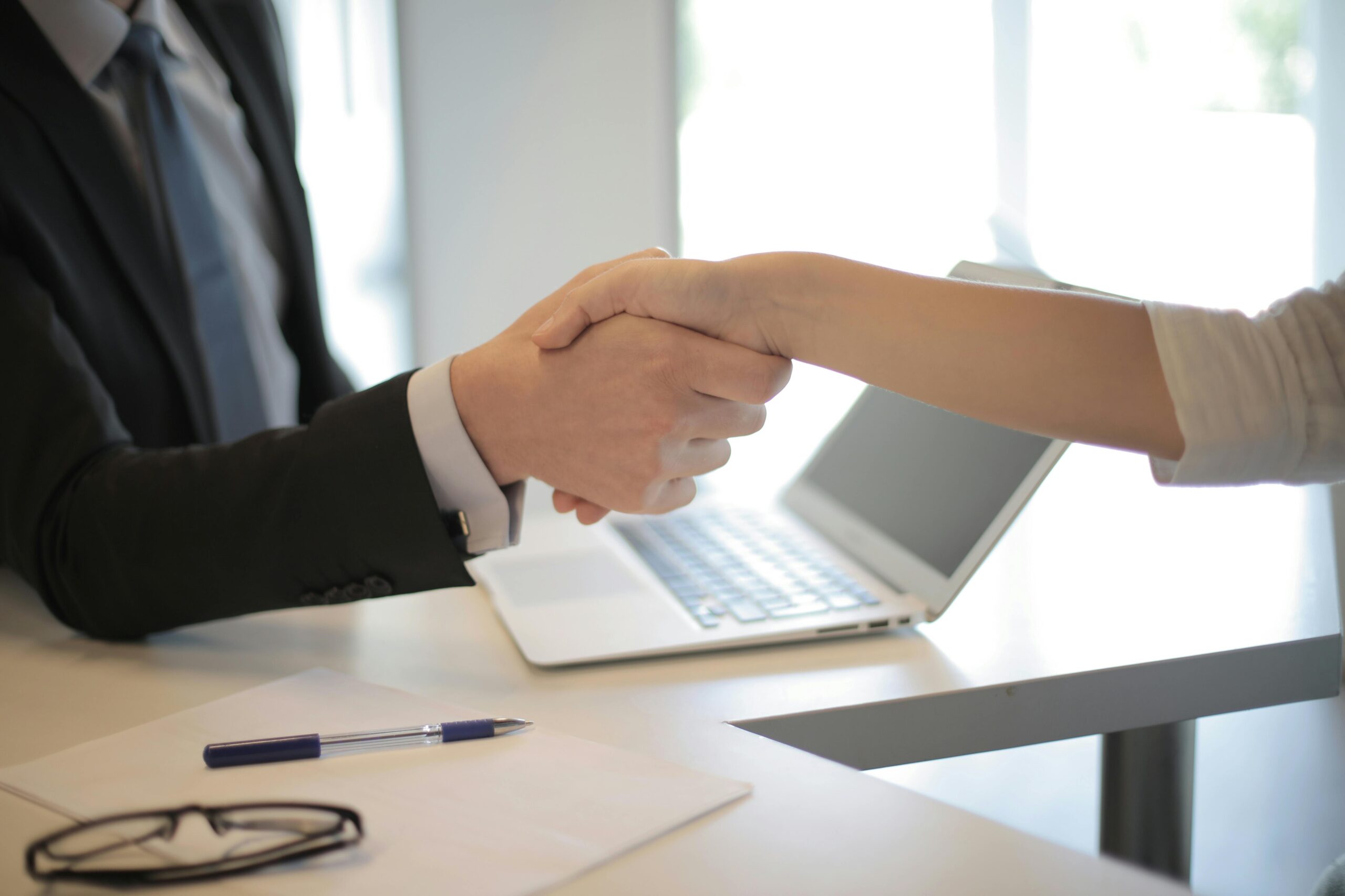 A close up of the hands of two people in office attire performing a handshake with a desk and laptop in the background.